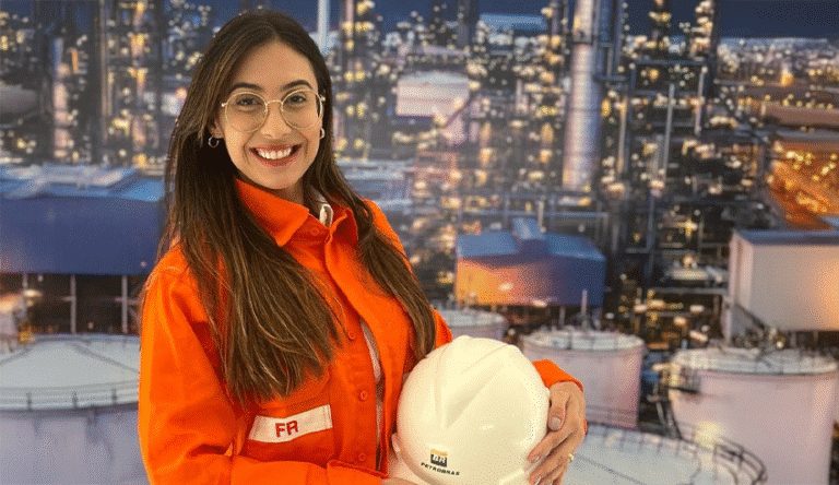 Jovem engenheira sorridente usando óculos e uniforme laranja da Petrobras, segurando capacete branco com logo da empresa, em frente a instalação industrial petrolífera iluminada